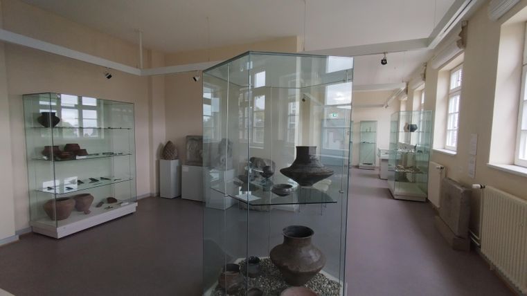 Ein Museum mit Vitrinen, die verschiedene Tongefäße und archäologische Artefakte ausstellen. Der Raum ist hell und hat große Fenster.