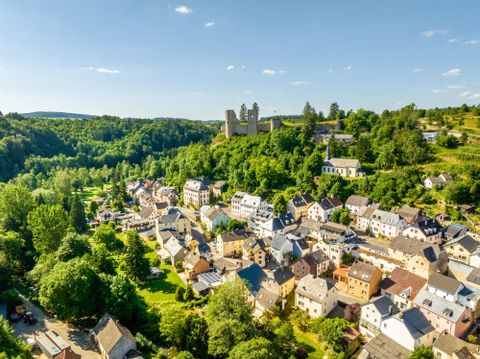 A picturesque town surrounded by green hills and trees. In the background, an old castle rises above the rooftops of the houses.