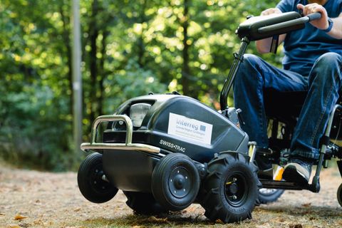 Un appareil motorisé pour l'extérieur, fixé à un fauteuil roulant. Il est utilisé sur un terrain accidenté dans la forêt.