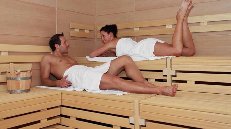 Ein Paar entspannt in einer Sauna auf Holzbank. Sie tragen Handtücher und genießen die warme Atmosphäre.