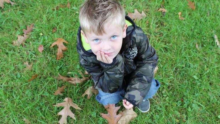 Ein kleiner Junge sitzt auf dem Gras mit nachdenklichem Ausdruck. Um ihn herum liegen einige Herbstblätter.