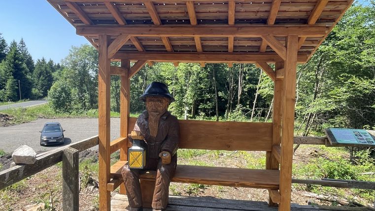 Un banc en bois avec une statue d'un homme tenant une lanterne. En arrière-plan, on peut voir des arbres et une rue.