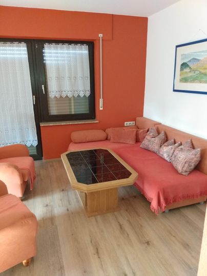 Ein gemütlicher Wohnbereich mit einem orangefarbenen Sofa und einem Holzkaffee Tisch. Große Fenster lassen viel Tageslicht herein.