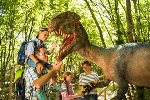 Une famille se tient dans une forêt et interagit avec un modèle de dinosaure. Les enfants sont ravis, tandis que les adultes sourient et jouent avec le dinosaure.