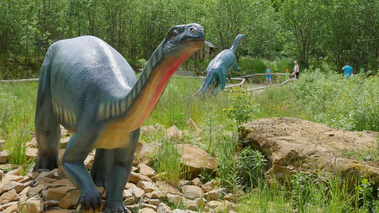 Deux modèles de dinosaures se tiennent dans un parc vert. Le paysage est adorné d'arbres et de pierres.