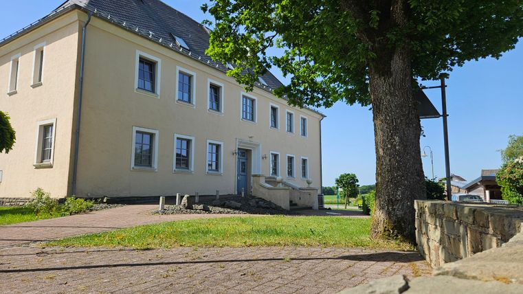 An old building with a yellow facade and several windows stands in a quiet environment. A large tree is visible in the foreground.