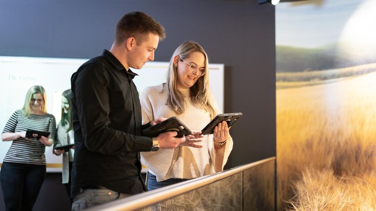 Twee personen staan in een kamer en kijken naar tablets. Op de achtergrond is een derde persoon te zien die ook een tablet gebruikt.