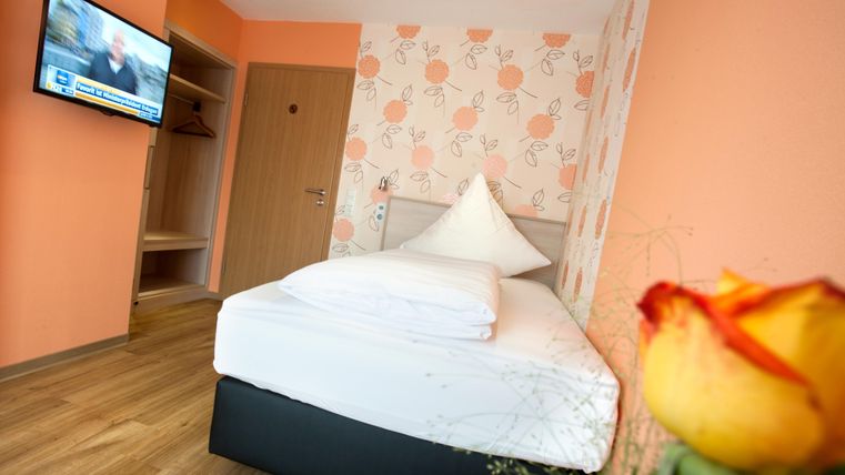 Ein gemütliches Zimmer mit einem Einzelbett und modernen Annehmlichkeiten. An der Wand ist ein Fernseher und die Wände sind freundlich in einem Orangeton gestaltet.