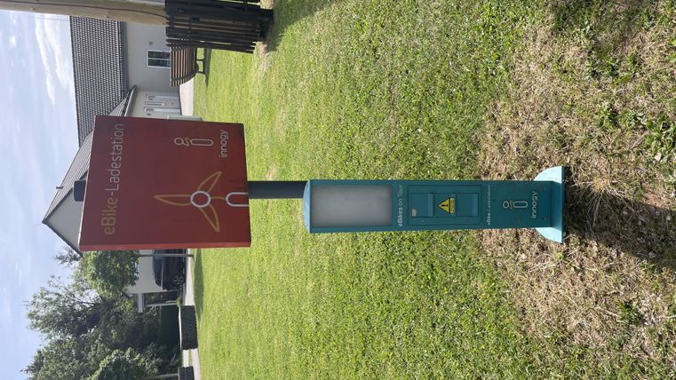 Eine Informationssäule steht auf einer grünen Rasenfläche. Im Hintergrund sind Häuser und ein blauer Himmel zu sehen.