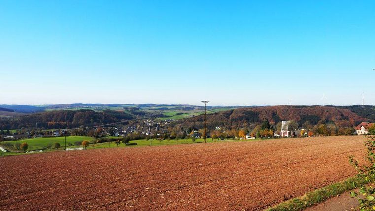 Eine weite Landschaft mit einem Acker im Vordergrund und sanften Hügeln im Hintergrund. Der Himmel ist klar und blau, ideal für einen sonnigen Tag.