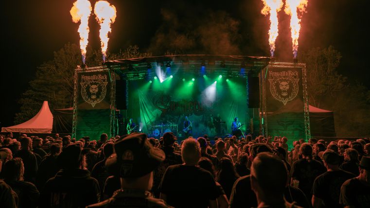 A concert stage with green lights and fire effects. Many spectators enjoy the music under the night sky.