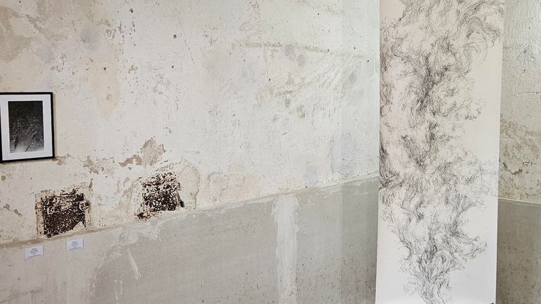 Ein leerer Raum mit grauen Wänden und einem Betonboden. Auf der linken Seite hängt ein kleines Bild, während sich auf der rechten Seite eine große, gemusterte Oberfläche befindet.