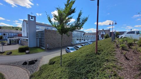 A modern building with large windows and a well-maintained environment. In the foreground, there is a green hill with bushes and a clear blue sky.