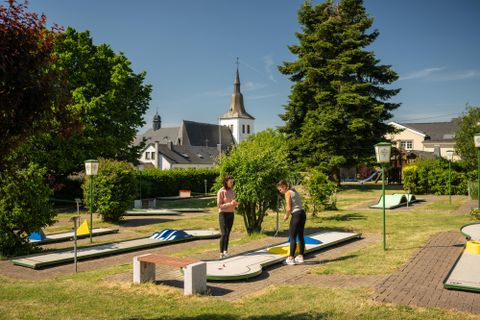 Eine Minigolfanlage mit mehreren Bahnen und Grünflächen. Im Hintergrund ist eine Kirche und blauer Himmel zu sehen.