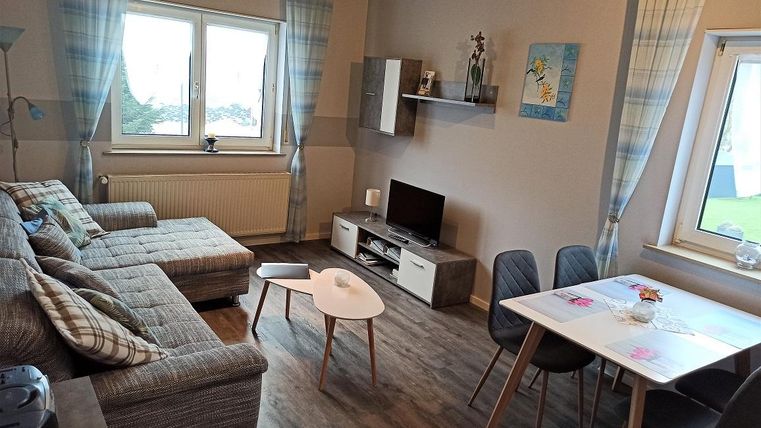 Ein gemütliches Wohnzimmer mit einem bequemen Sofa und einem kleinen Tisch. Es gibt eine TV-Einheit und einen Essbereich mit Stühlen am Fenster.