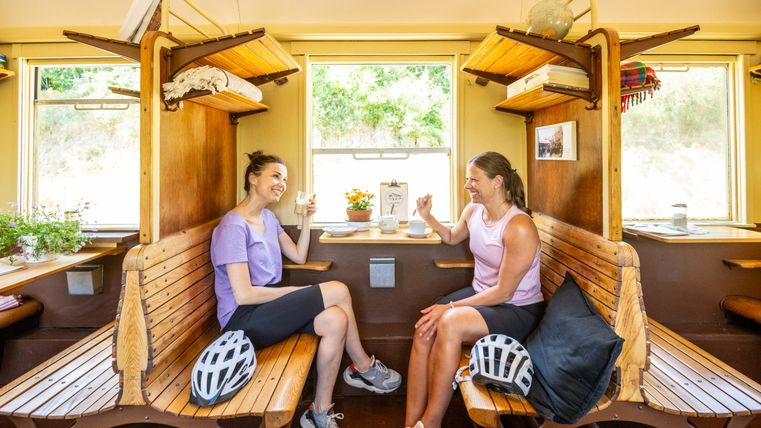 Zwei Frauen sitzen in einem hellen Raum auf Holzbank. Sie genießen Getränke und lachen miteinander.