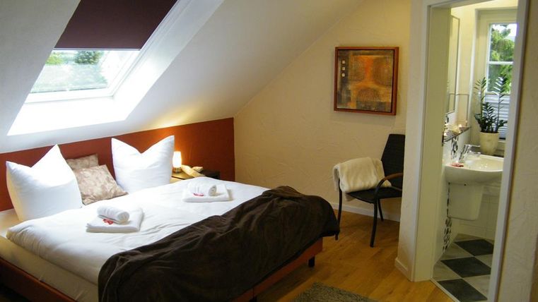 Gemütliches Schlafzimmer mit einem großen Bett, weißen Kissen und moderner Einrichtung. Der Raum hat ein Fenster mit natürlichem Licht und führt zu einem Badezimmer.