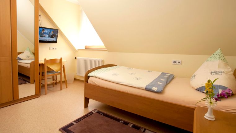 Ein einfaches Zimmer mit einem Einzelbett, einem Fernseher und einem Spiegel. Die Wände sind in hellem Farbton gestrichen und es gibt eine kleine Vase mit Blumen auf dem Tisch.