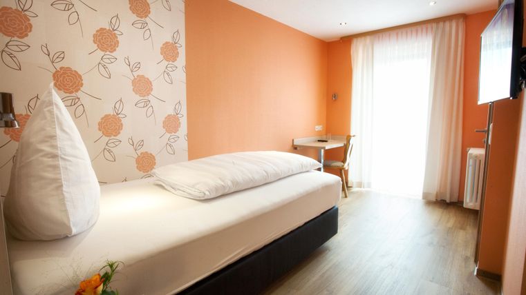 Ein gemütliches Einzelzimmer in sanften Orangetönen. Es verfügt über ein Bett, einen Tisch und einen Zugang zum Balkon mit viel natürlichem Licht.