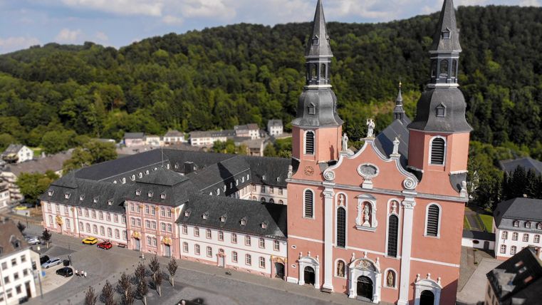 Eine beeindruckende Kirche mit zwei Türmen in einer malerischen Umgebung. Im Vordergrund befindet sich ein Platz mit Bäumen und historischen Gebäuden.
