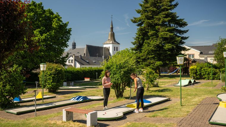 Eine Minigolfanlage mit mehreren Bahnen und Grünflächen. Im Hintergrund ist eine Kirche und blauer Himmel zu sehen.