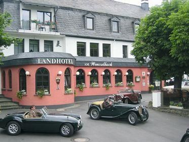 Ein charmantes Landhotel mit rosa Fassade und grünen Bäumen. Vor dem Hotel parken klassische Autos.