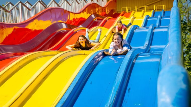 Trois enfants glissent joyeusement sur les toboggans colorés. Le ciel ensoleillé et les couleurs vives rendent la scène accueillante et ludique.