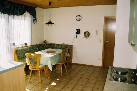 Eine gemütliche Küche mit einem Esstisch und einer Eckbank. Die Wände sind hell, und es gibt eine Fensterfront mit Vorhängen.