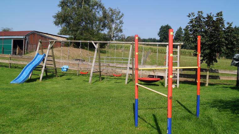 Ein Spielplatz mit einer Rutsche, Klettergerüst und Schaukeln auf grünem Rasen. Im Hintergrund sind Bäume und ein Gebäude sichtbar.