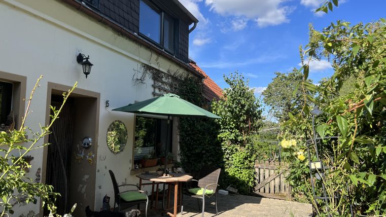 Ein einladender Außenbereich mit einem Tisch und Stühlen unter einem grünen Sonnenschirm. Im Hintergrund sieht man einen gepflegten Garten und einen blauen Himmel mit einigen Wolken.