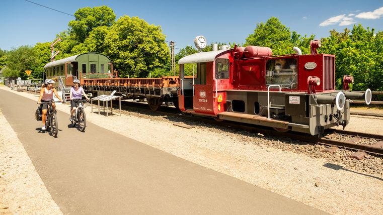 Eine rote Lokomotive steht neben den Gleisen, während zwei Radfahrer auf einem Weg vorbeifahren. Der Himmel ist blau und die Umgebung ist grün und sonnig.