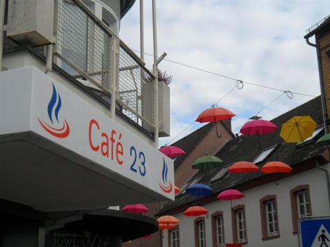 Ein gemütliches Café mit dem Namen „Café 23“ ist zu sehen. Bunte Regenschirme hängen dekorativ über der Straße.