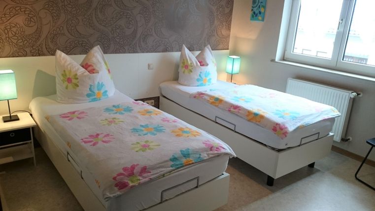 Ein gemütliches Schlafzimmer mit zwei Einzelbetten in hellem Design. Die Betten sind mit bunten Bettdecken dekoriert und es gibt Lampen und Fenster für natürliches Licht.