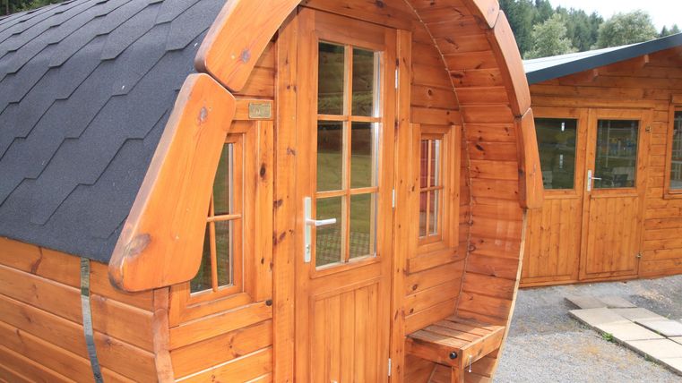 Ein hölzernes, rundes Häuschen mit einem großen Fenster und einer Eingangstür. Im Hintergrund sind weitere ähnliche Gebäude zu sehen.