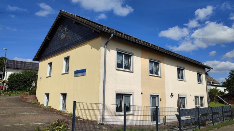 Ein zweigeschossiges Wohnhaus mit einer hellen Fassade. Es steht in einer ruhigen Umgebung unter einem blauen Himmel.