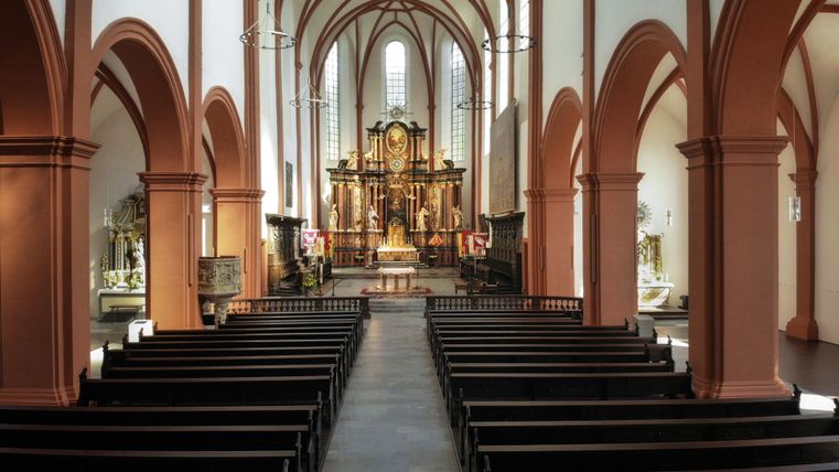 Eine Kirche mit bunten Fenstern und einem schönen Altar im Hintergrund. Die Bänke sind ordentlich aufgereiht und die Wände sind in warmen Farbtönen gehalten.
