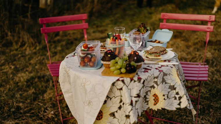 Ein gemütlicher Tisch im Freien mit bunten Stühlen. Auf dem Tisch stehen verschiedene Snacks, Obst und Getränke auf einer hübschen Tischdecke.