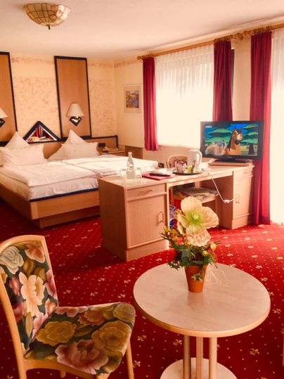 Ein gemütliches Hotelzimmer mit einem großen Bett, einem Fernseher und einem kleinen Tisch. Die Möbel sind hell und der Teppich hat ein rotes Muster.