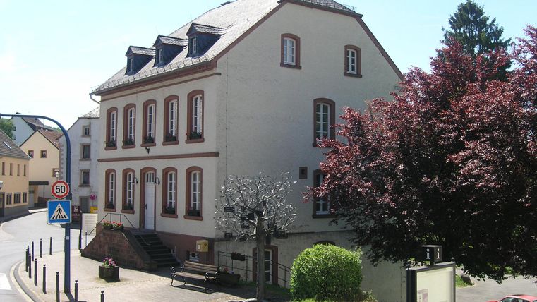 A building with several windows and a beautiful garden. Trees and shrubs surround the corner of the street.