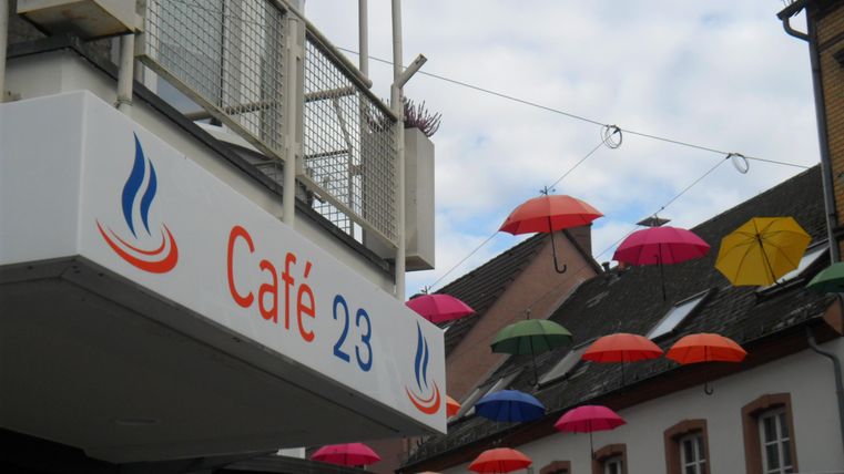 Ein gemütliches Café mit dem Namen „Café 23“ ist zu sehen. Bunte Regenschirme hängen dekorativ über der Straße.