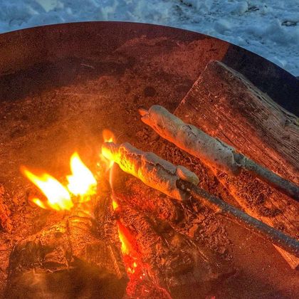 Ein gemütliches Lagerfeuer mit zwei Stöcken, an denen Teig gebacken wird. Die Flammen leuchten orange und die Umgebung ist schneebedeckt.