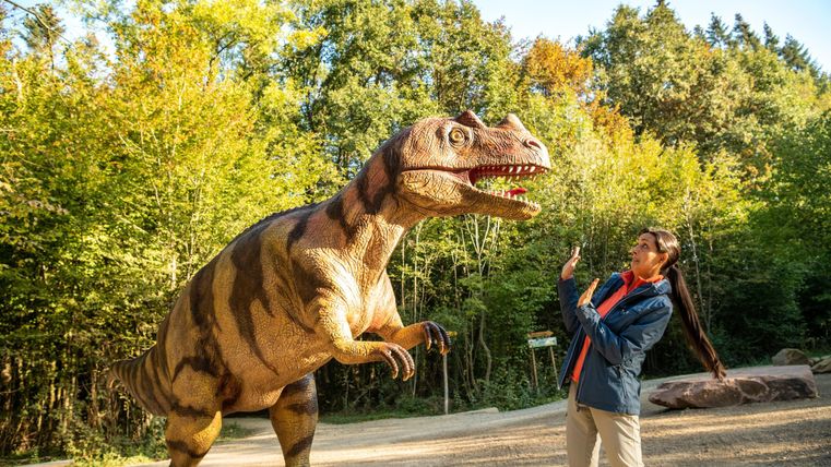 Une personne se tient devant un grand modèle de dinosaure dans la forêt. Le soleil brille et l'environnement est vert et naturel.