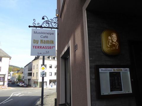 Ein Schild für eine Musikkneipe und Café mit dem Namen "by Namik" ist an der Wand angebracht. Es ist eine ruhige Straße mit mehreren Gebäuden im Hintergrund.