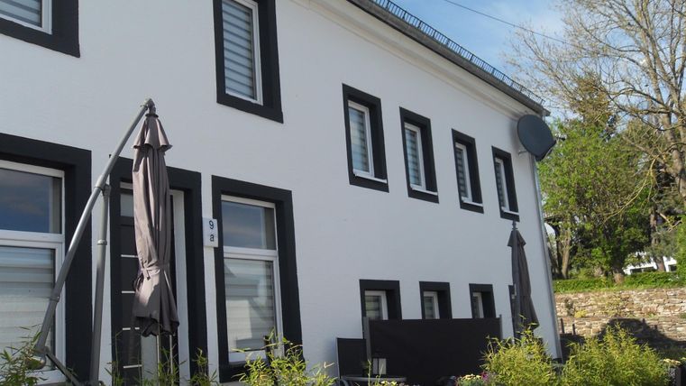 Ein modernes, zweigeschossiges Haus mit weißen Wänden und schwarzen Fensterrahmen. Vor dem Gebäude stehen Pflanzkübel mit grünen Pflanzen.