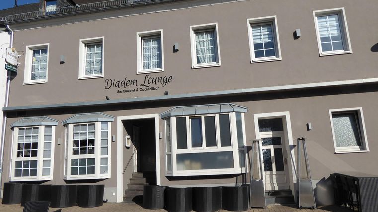 Ein modernes Restaurant namens Diadem Lounge mit großen Fenstern und einem einladenden Eingangsbereich. Die Fassade ist in einem hellen Grauton gehalten und die Umgebung wirkt ordentlich und gepflegt.