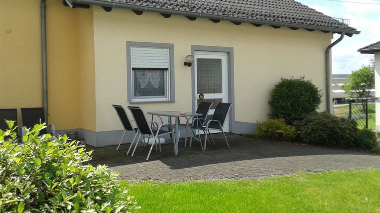 Ein gemütlicher Sitzbereich mit einem Tisch und Stühlen steht vor einem gelben Haus. Die grüne Rasenfläche und die Pflanzen schaffen eine einladende Atmosphäre.