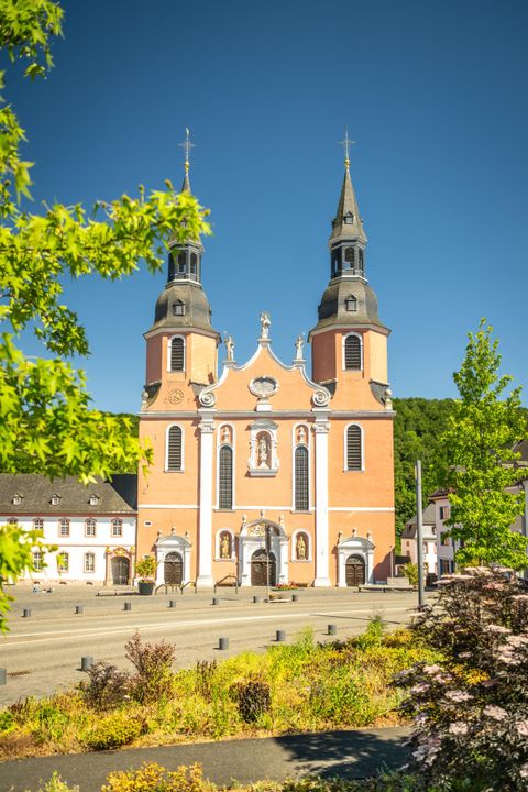 Eine beeindruckende Kirche mit zwei hohen Türmen und einer schönen Fassade in hellen Farben. Umgeben von Bäumen und einem gut gestalteten Platz.
