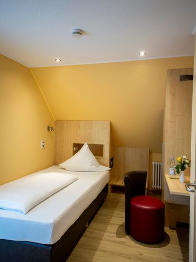 Ein gemütliches Zimmer mit einem Einzelbett, einem roten Hocker und einem Schreibtisch. Die Wände sind in einem warmen Gelbton gehalten, was eine einladende Atmosphäre schafft.