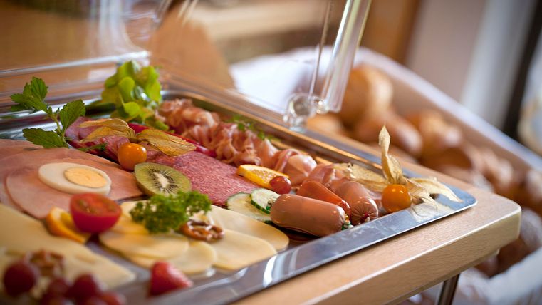 Eine ansprechend dekorierte Platte mit verschiedenen Wurst- und Käsesorten, frischem Obst und Gemüse. Im Hintergrund sind Brötchen sichtbar, die das Frühstücksbuffet ergänzen.