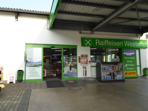 Ein Geschäftseingang mit großen Fenstern und grünen Akzenten. Es gibt ein Sortiment an Snacks und Getränken sowie eine Lotto-Annahmestelle.